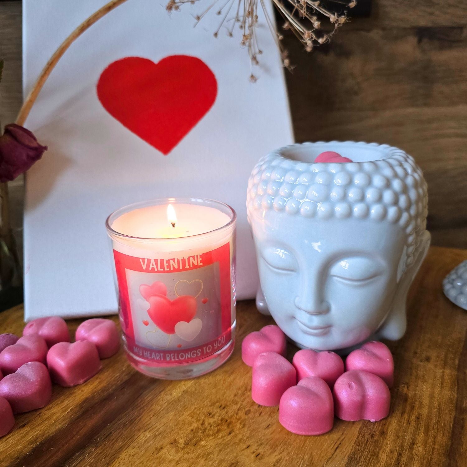 a lit valentine candle in a glass container with red heart design surrounded by wax melt hearts and a buddha head wax melter