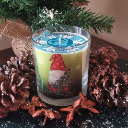 Father Christmas candle with father christmas holding a christmas tree design on the front of a scented candle in glass container. The candle is surrounded by pine cones.
