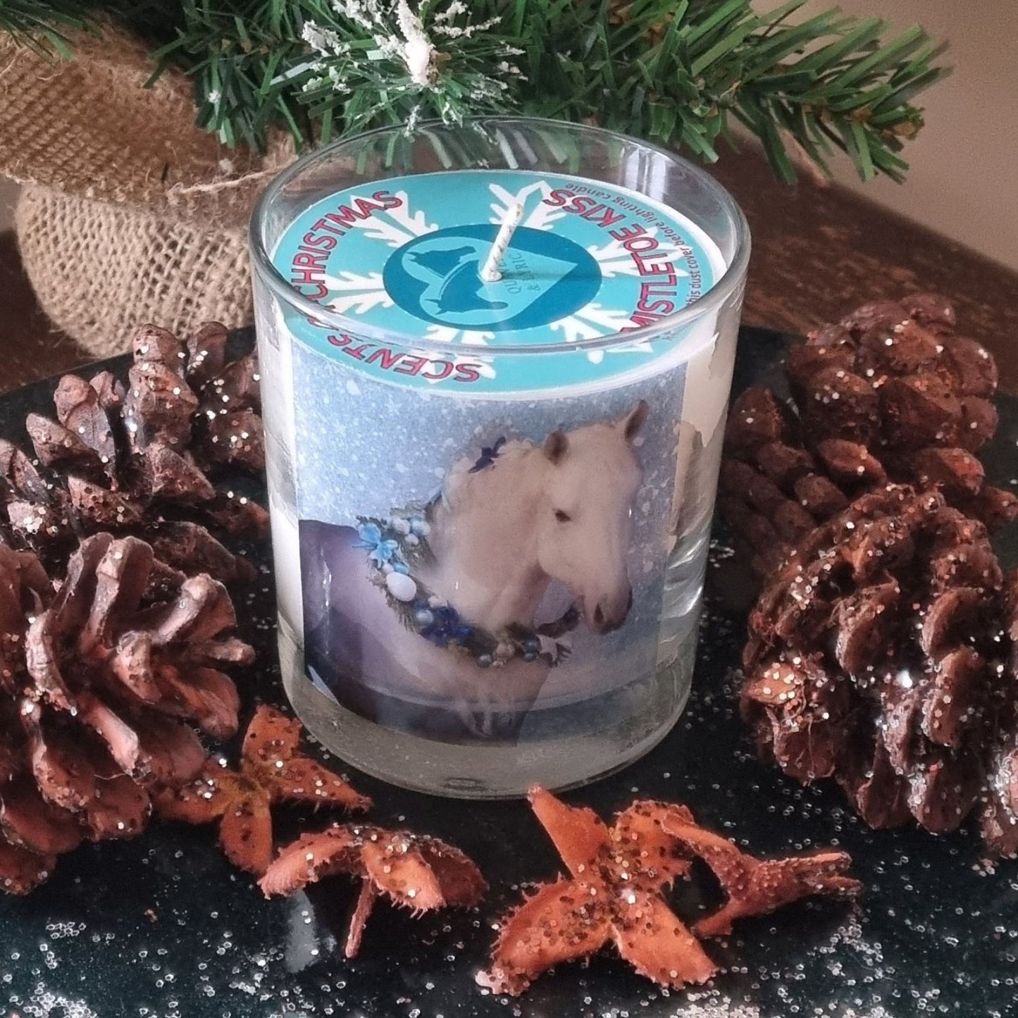 a scented candle in a glass container with a grey horse wearing a festive garland on his neck. the candle is surrounded with glittery pine cones.