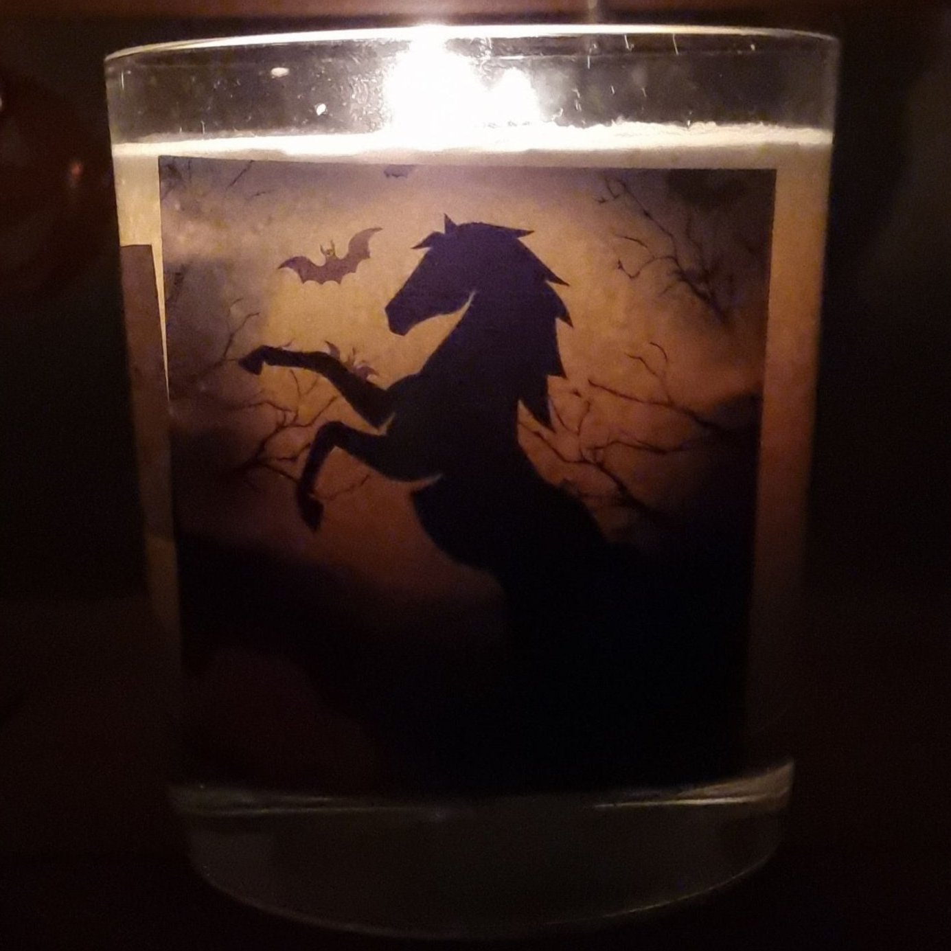 a lit scented halloween candle in a glass container with a black rearing horse and pumpkins in a graveyard setting