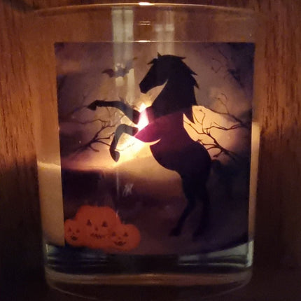a lit scented halloween candle in a glass container with a black rearing horse and pumpkins in a graveyard setting