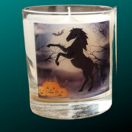 a scented halloween candle in a glass container with a black rearing horse and pumpkins in a graveyard setting