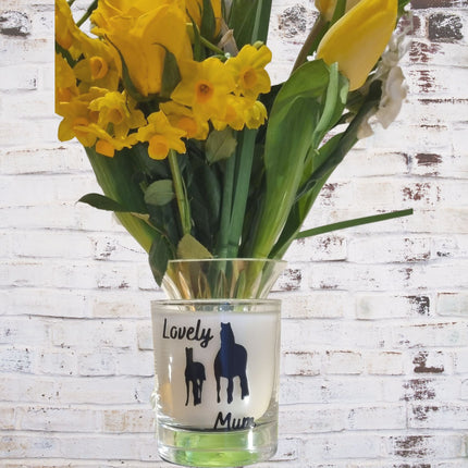 A scented candle in a glass container from Quaver&Lyric, featuring a Lovely Mum horse design, is displayed against a white brick wall—perfect as a Mothers Day gift.