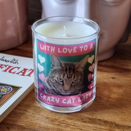 The Quaver&Lyric Scented Candle In Glass Container Crazy Cat Lady Bengal Cat Design features a tabby cat label with the message With love to a crazy cat lady and colorful hearts, placed on wood beside books, a pink vase, and gift wrap.