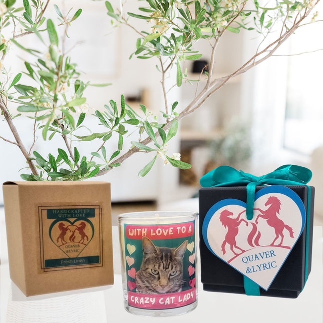 A Scented Candle In Glass Container Crazy Cat Lady Bengal Cat Design by Quaver&Lyric rests between a handcrafted brown box and a black gift box with blue ribbon, gift wrap, and a heart tag reading Quaver&Lyric.