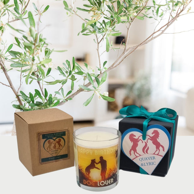 A Quaver&Lyric Scented Candle In Glass Container labeled Happy Birthday Dog Lover sits by its brown box. Beside it, a heart-shaped dog ornament with Quaver&Lyric is on a black gift box with teal ribbon and a leafy branch behind.