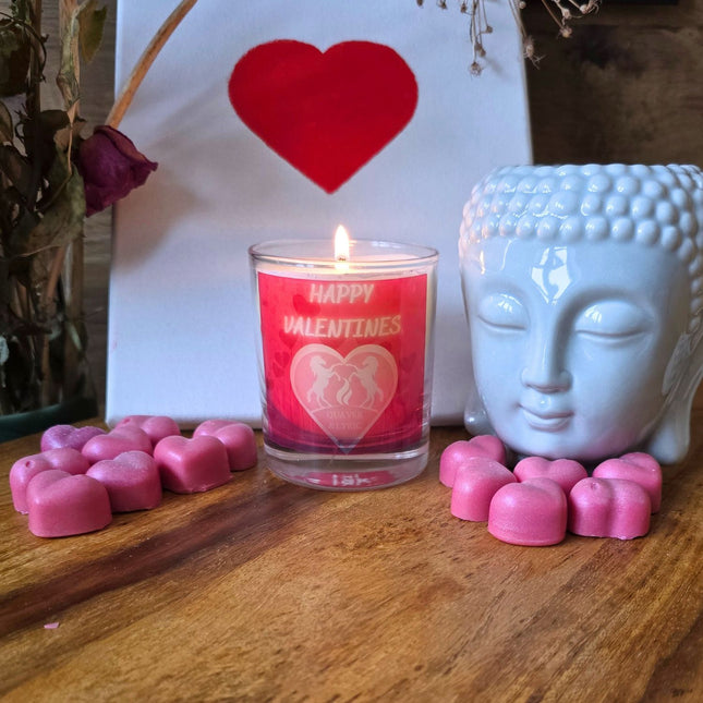 The Quaver&Lyric Scented Happy Valentine Candle (30cl) in a glass container sits on wood, surrounded by pink heart chocolates, a white Buddha head planter, fragrances, and a canvas with a red heartâ€”handmade in Barton Seagrave.
