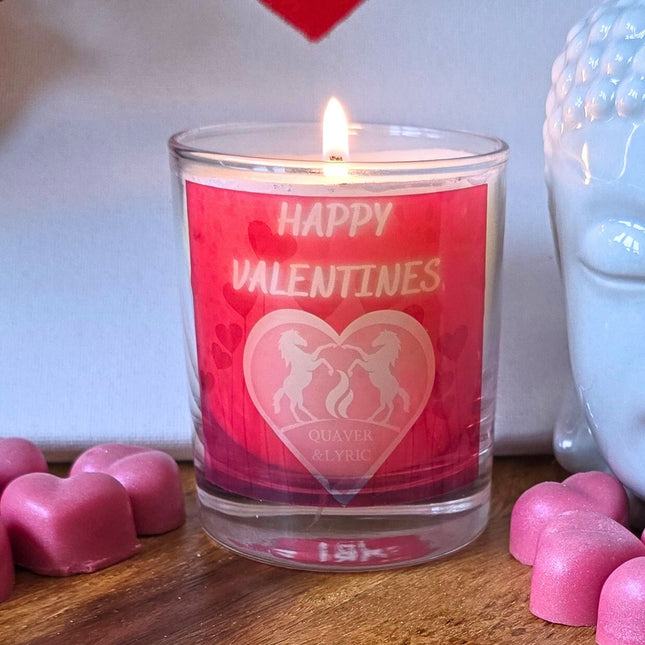 The Quaver&Lyric Scented Happy Valentine Candle In Glass Container (30cl) features a pink HAPPY VALENTINES label with two unicorns inside a heart and is shown with pink hearts and part of a white Buddha statue nearby.