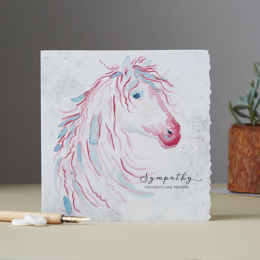 a square greeting card with a crinkled edge on one side resting on a surface with an ink pen and polos in front and a planr behind. The card features a beautiful horse with a flowing mane in pink and blue grey with wording on the outside Sympathy, Thoughts and Prayers