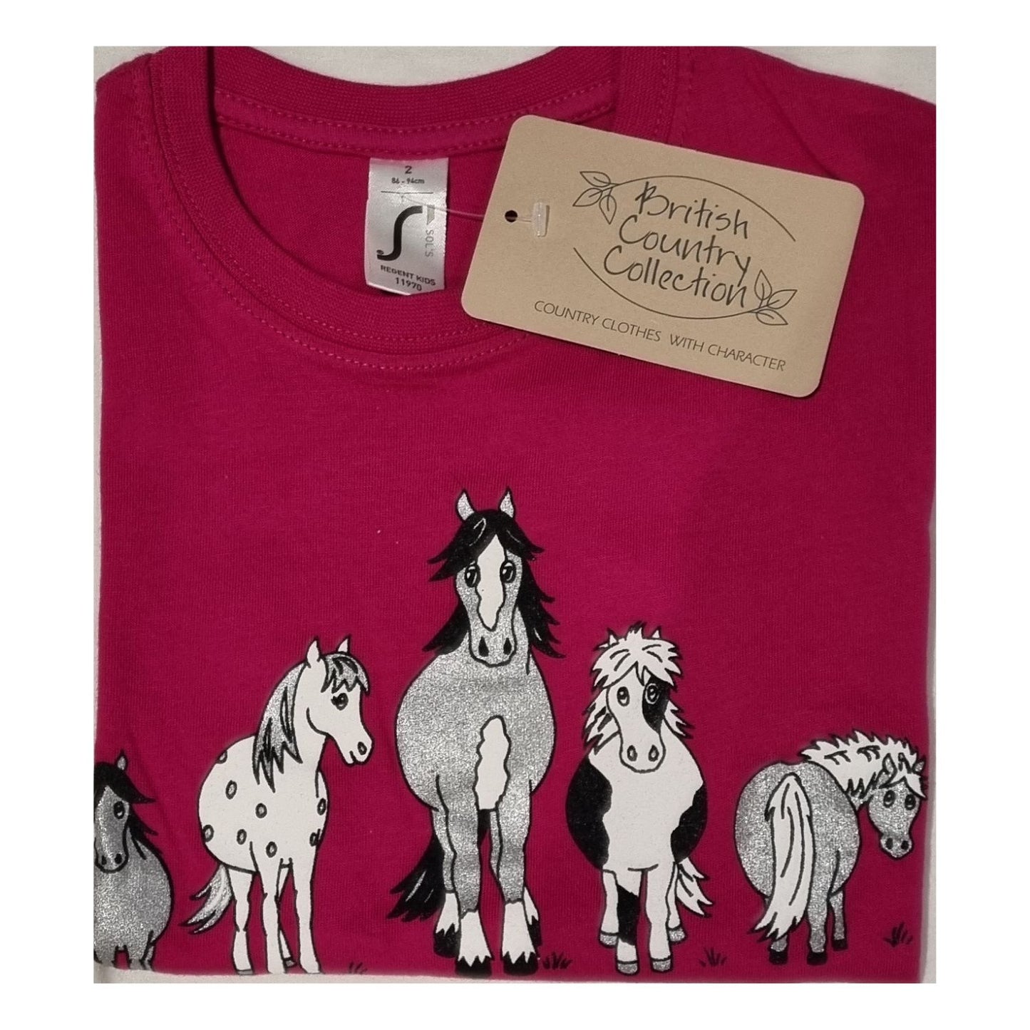 The Childrens T Shirt Printed The Herd Horse Design by British Country Collection is a folded pink 100% cotton tee featuring five uniquely patterned cartoon horses and a beige brand tag. Machine washable for easy care.