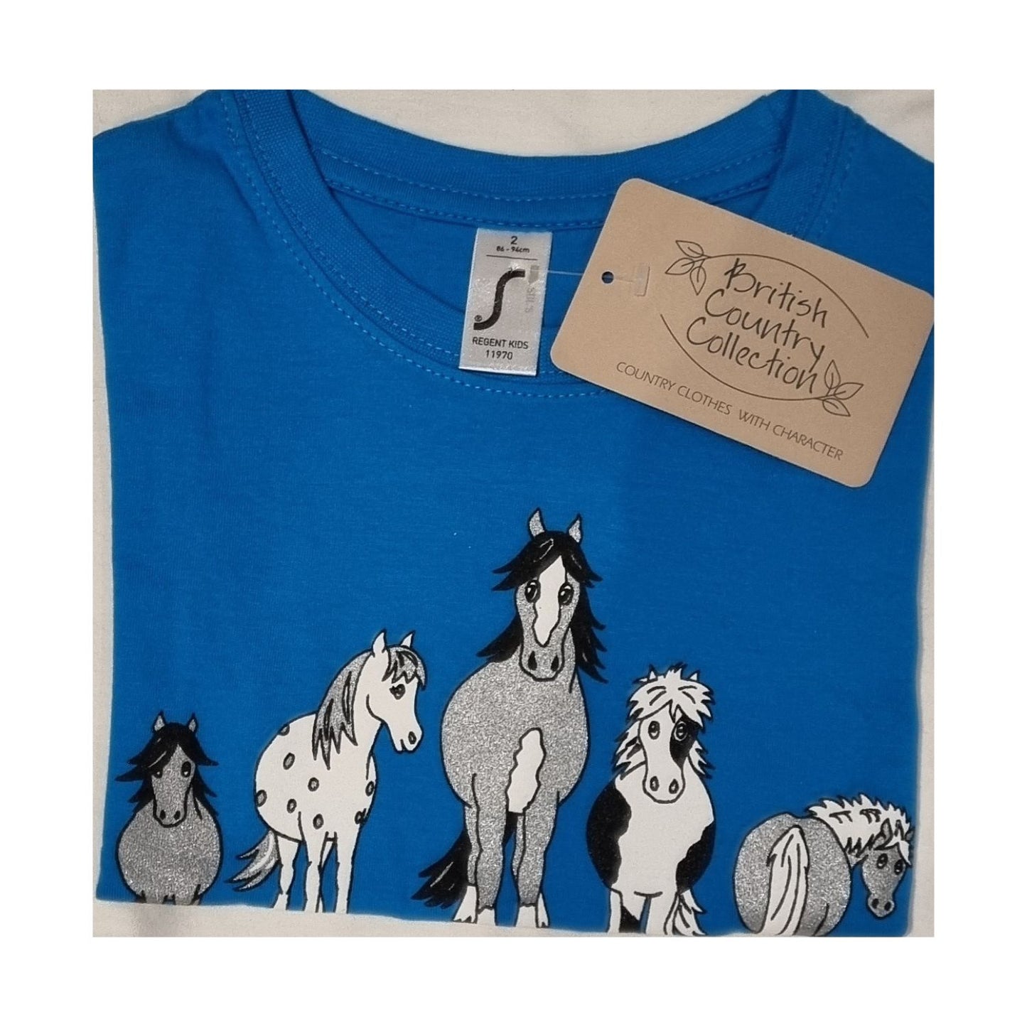 A folded blue Childrens T Shirt Printed The Herd Horse Design by British Country Collection, made from 100% cotton, features cartoon horses on the front and a branded tag attached to the collar.
