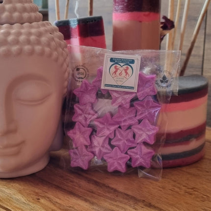 A ceramic Buddha head sits next to a Quaver&Lyric Pack of 15 Soy Wax Melt Stars. Behind them, colorful candles and wooden reeds on a wooden table create a serene, fragrance-filled atmosphere.