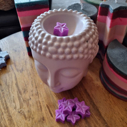 A Quaver&Lyric Buddha head bowl on a wooden table displays a purple star from the Soy Wax Melt Stars Quaver&Lyric Pack of 15, with more fragrant melts scattered nearby and colorful handmade decor in the background.