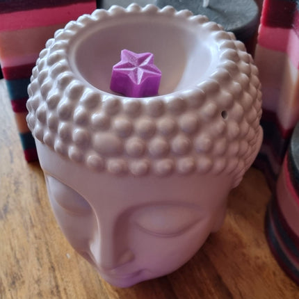 A ceramic Buddha head candle holder displays a handmade purple soy wax star from the Quaver&Lyric Soy Wax Melt Stars Pack of 15, set on wood with colorful candles behind and a soothing fragrance in the air.