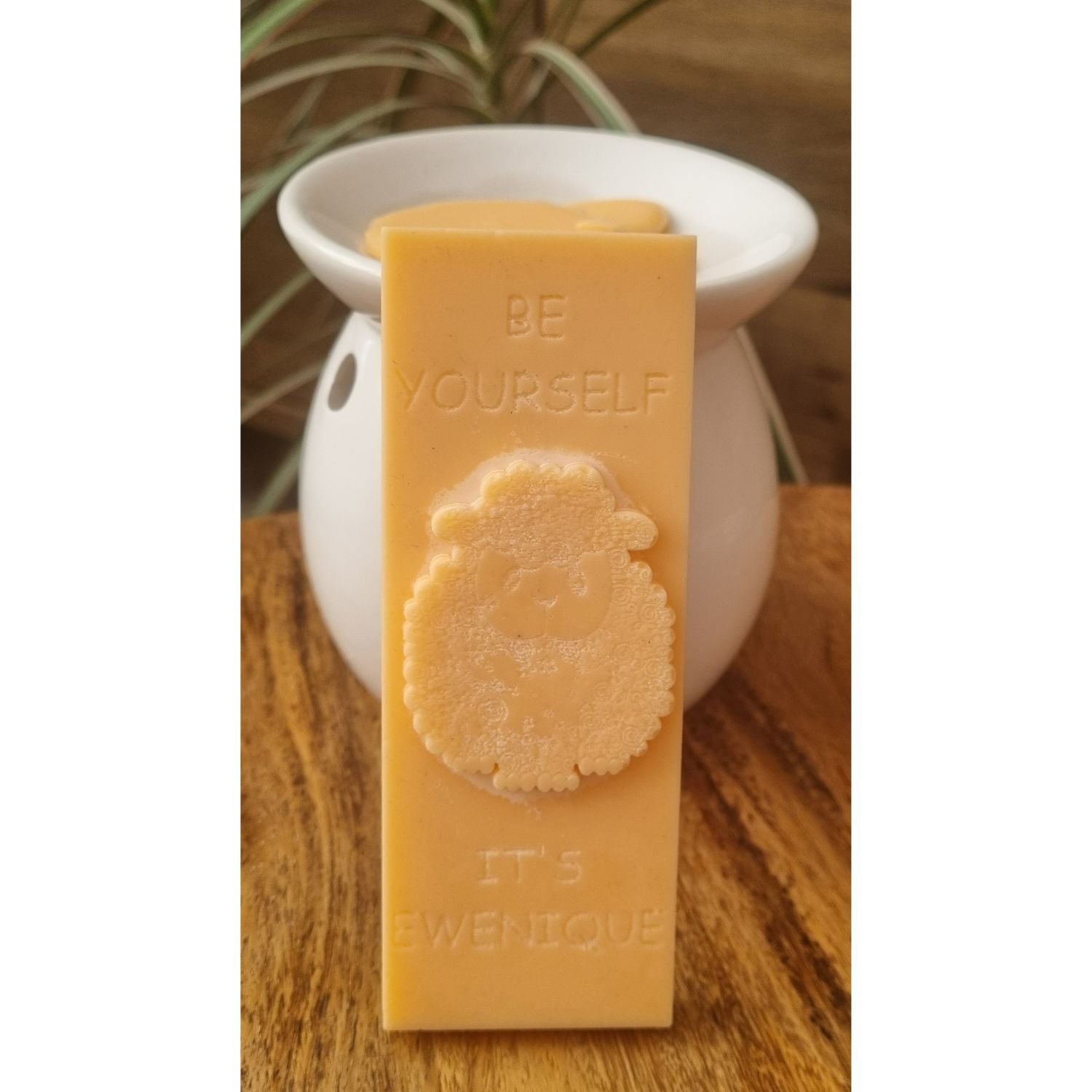 An orange coloured rectangle of wax with a raised sheep design with the writing be yourself above and it's ewenique below. The bar is leaning against a white ceramic wax melter on a wooden table.