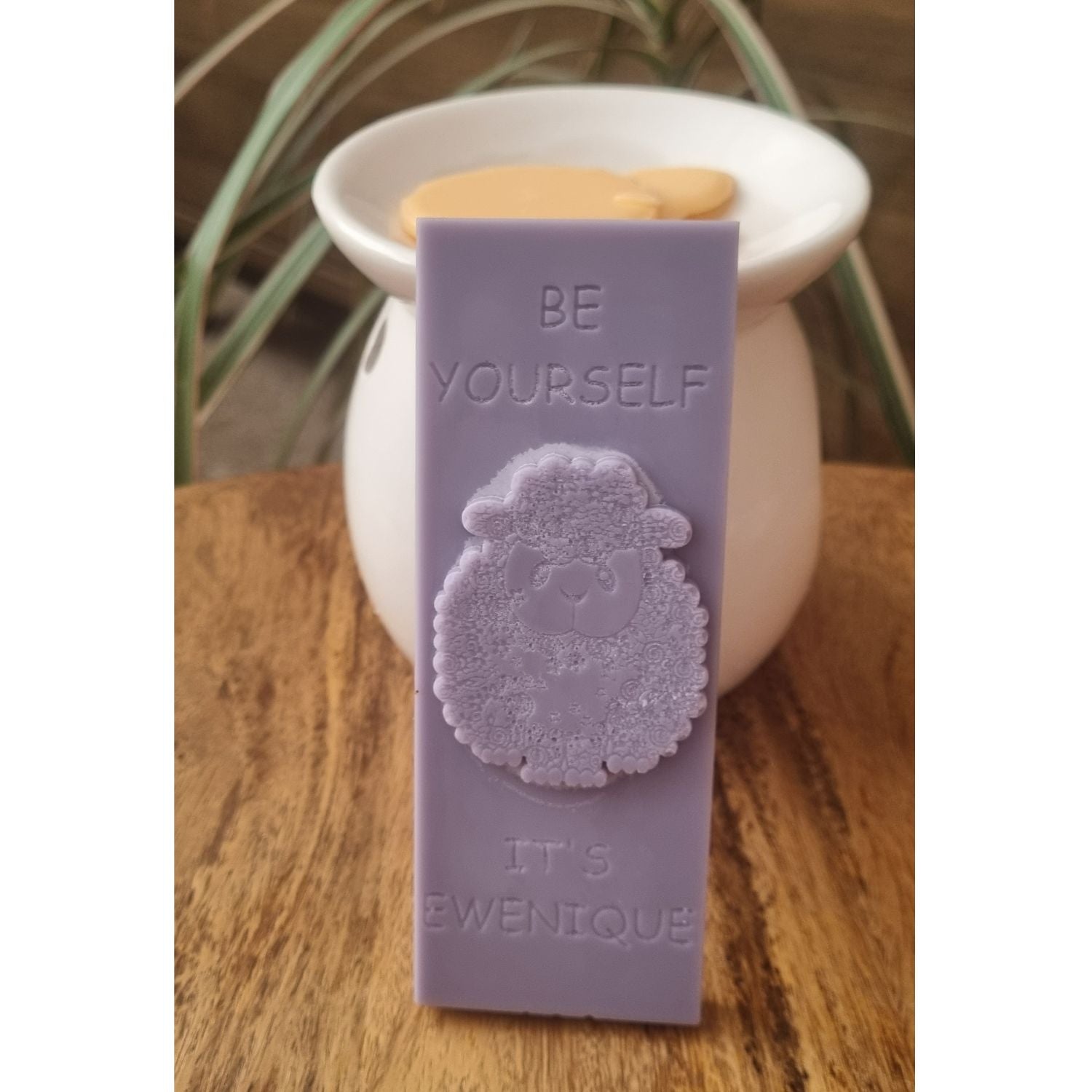 A lilac coloured wax melt bar with a raised sheep design with the writing be yourself above and it's ewenique below next to a wax warmer