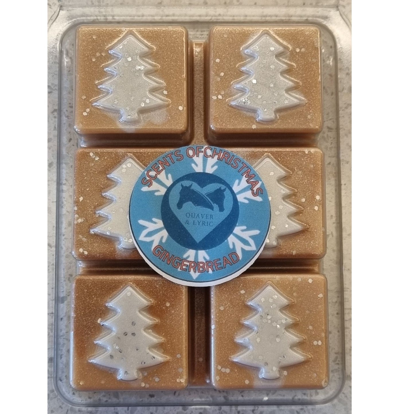 Christmas fragrance wax melts in a Christmas tree design clamshell. With white scented wax christmas trees on golden brown scented cubes. With  Quaver and Lyric branded label.