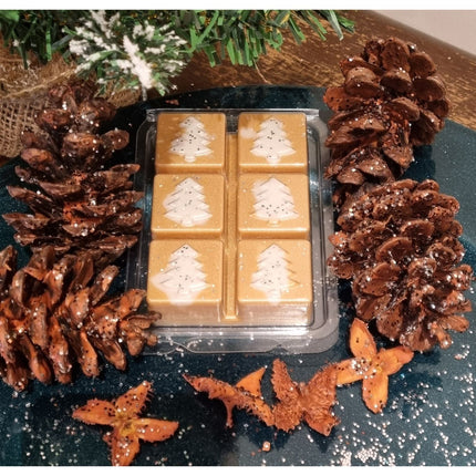 Christmas fragrance wax melts in a Christmas tree design clamshell. With white scented wax christmas trees on golden  brown scented cubes. In a festive setting with pine cones.