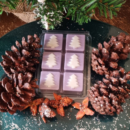 Christmas fragrance wax melts in a Christmas tree design clamshell. With white scented wax christmas trees on lilac coloured scented cubes. In a festive setting with pine cones.