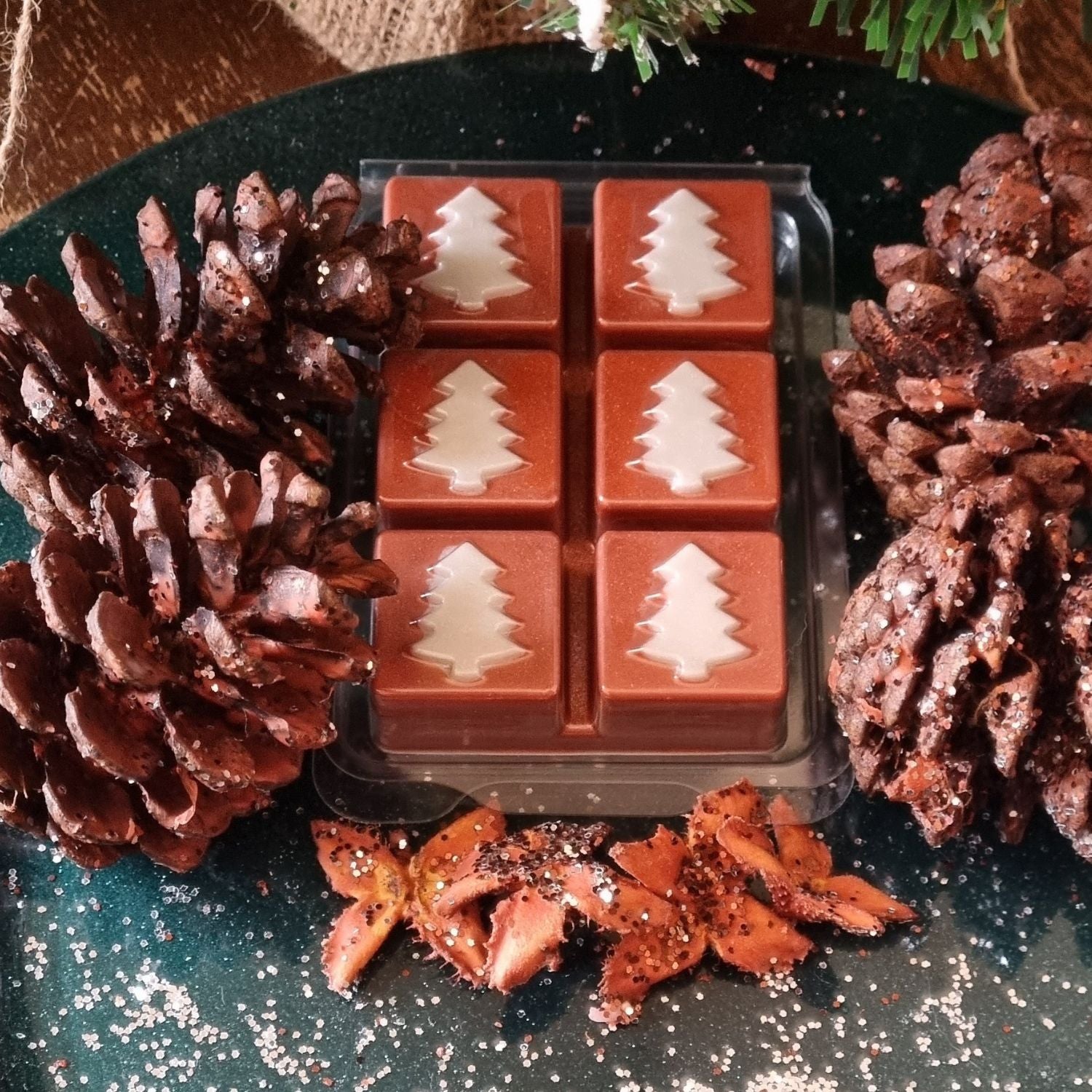 Christmas fragrance wax melts in a Christmas tree design clamshell. With white scented wax christmas trees on nut brown scented cubes. In a festive setting with pine cones.